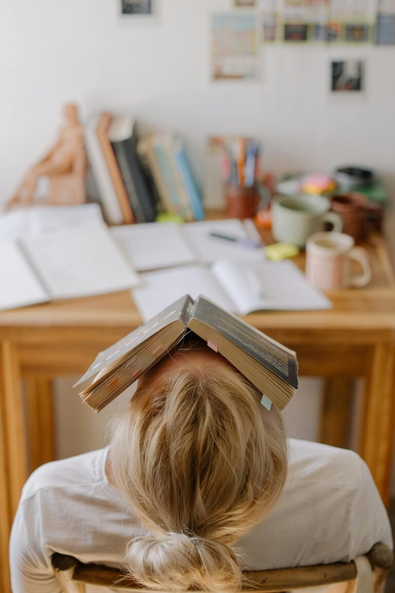 Iemand zit met een opengeslagen boek op het hoofd aan een bureau vol studiemateriaal