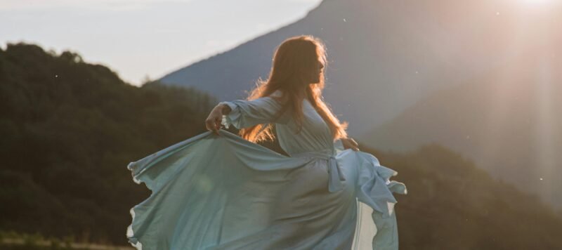 Vrouw in een wapperende jurk danst in een veld voor een berg bij zonsondergang