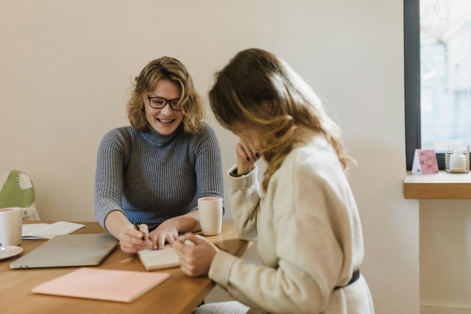 Twee vrouwen in gesprek aan een tafel, een van hen maakt aantekeningen tijdens een coachsessie bij daglicht
