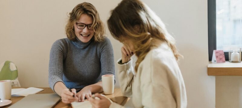 Twee vrouwen in gesprek aan een tafel, een van hen maakt aantekeningen tijdens een coachsessie bij daglicht