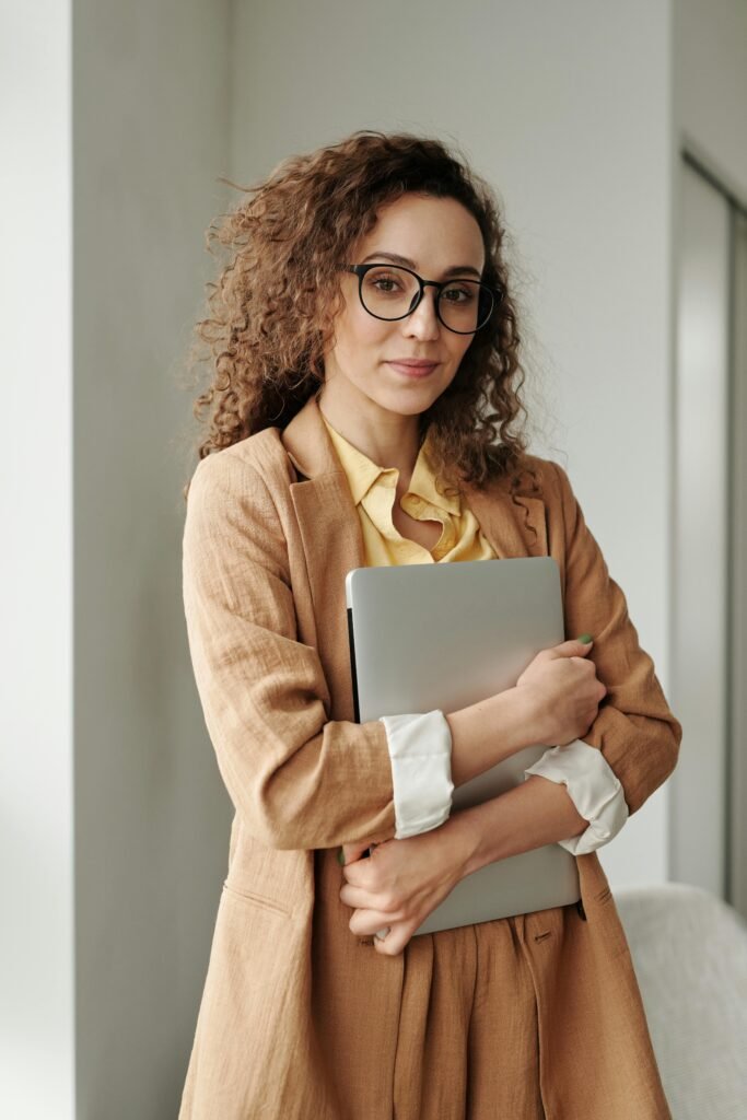 Vrouw met bril en beige colbert houdt een laptop tegen zich aan