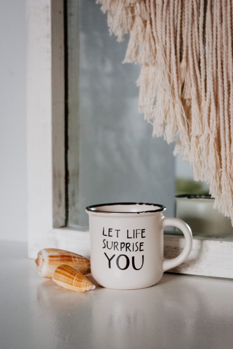 Mok met de tekst let life surprise you op een vensterbank naast twee schelpen