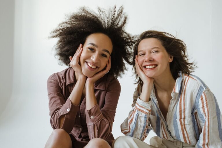 Twee vrouwen zitten lachend naast elkaar tegen een lichte muur