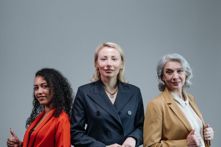Drie vrouwen in gekleurde blazers staan naast elkaar tegen een grijze wand