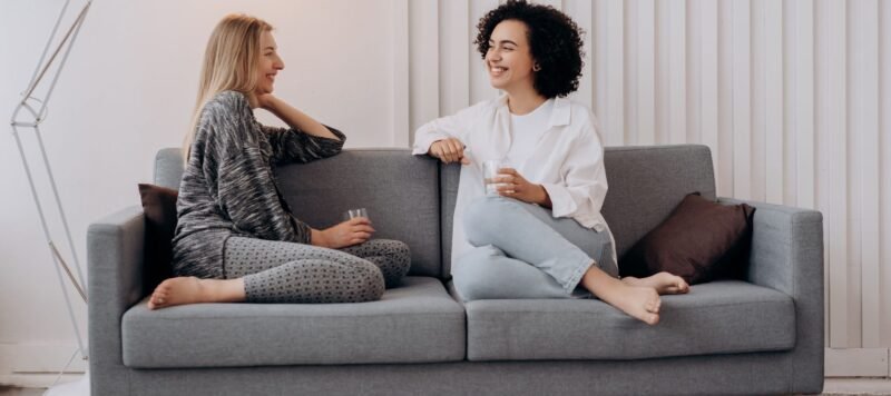 Twee vrouwen zitten ontspannen in gesprek op een bank in een lichte woonkamer