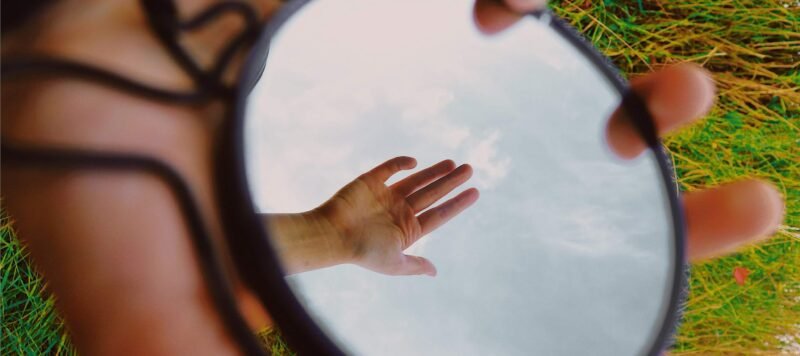 Ronde spiegel in het gras waarin een uitgestrekte hand en de lucht weerspiegelen